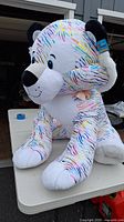 Full view of the very large multicolored striped bear plush, white with colorful stripes, sitting on a table.