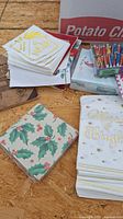 Photo showing stack of various holiday-themed paper napkins including holly leaf, Christmas tree, and 'Merry & Bright' designs, some plain white with gold accents, placed on a wooden surface with a box labeled 'Potato Chips' in the background and colorful birthday candles nearby.