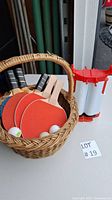 Five ping pong paddles with red and black paddle faces and light wooden handles inside a wicker basket, three white ping pong balls visible, and a red and white plastic table divider to the right.