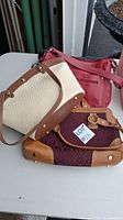 Three pocketbooks displayed on white surface, cream fabric with brown leather pocketbook on top, dark red leather handbag visible, brown fabric with logo print pocketbook at bottom.