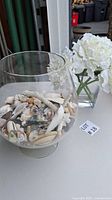 Full view of large clear glass bowl filled with assorted seashells set next to clear vase with white artificial flowers on a table.