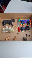 Photo of a cardboard box containing multiple small ceramic and resin animal figurines, including colorful horses and small animals with a red marker for size reference.
