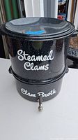 Black speckled enamel clam steamer pot labeled 'Steamed Clams' on top section and 'Clam Broth' on bottom section with a spout.