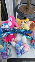 Photo showing a variety of colorful stuffed animals including bears, snakes with sequins, a penguin, and other plush toys piled on a surface.