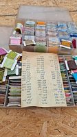 Top view of one open box showing multiple compartments with embroidery threads in various colors labeled with handwritten numbers on cardboard cards, plus a handwritten list of thread color codes.