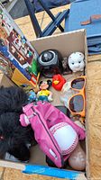 Top view of box showing various toys including LEGO set, plush dog in pink jacket, sunglasses, toy figurines, piggy bank, and black electronic device.