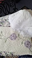 Quilted twin bedspread showing patterned embroidery of purple flowers and green leaves paired with a white cotton dust ruffle featuring delicate lace trim.