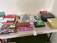 Overview of table with art supplies including paper pads, pastel sets, gouache tubes, storage tins, wooden box, pencils and pencil sharpener