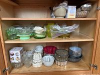 Three wooden shelves containing vintage glass juicers, assorted cake pans, molds, and boxed baking supplies