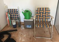 Two vintage folding lawn chairs, a clamp-on beach umbrella, a Coleman Party Stacker cooler, green watering can, and thermometers on floor.