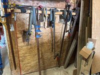 Full view of multiple clamps hanging on plywood wall including various sizes of C-clamps, bar clamps and quick grip clamps.
