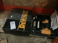 DeWalt branded black metal case and Bostitch brad nailer with yellow-orange body inside black case on garage floor.