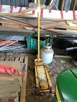 Yellow hydraulic floor jack positioned vertically with long handle, placed on a dirt floor near a propane tank and wood pieces.