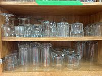 Wooden shelf displaying multiple clear glass items including snifters, wine glasses, tumblers, and other glassware.