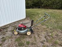 Side view of Honda push lawn mower showing metal silver deck, red engine housing, yellow wheel adjustment knobs, fabric clippings bag attached, and foldable handlebar.