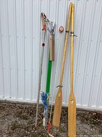 Full shot of two wooden oars and three long-handled pruning tools against a white metal wall.