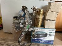 Multiple brass finish plant pots stacked, metal lizard wall decor piece resting on or near pots with a boxed item labeled Sony Poles