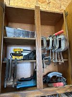 Wooden storage cabinet with tools arranged inside including Dewalt sander, electric power tool, and clamps.