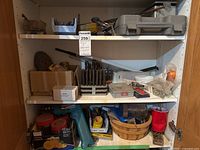 Three shelves of a white shelving unit filled with various tools and hardware including a Makita drill case, drill bits, and miscellaneous items.