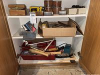 Cabinet shelf with multiple tools and hardware boxes, including wooden handled chisels and a Makita charger.