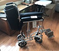 Side view of the folded wheelchair showing chrome aluminum frame, black fabric seat folded inward, footrests, and rear wheels.