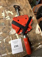 Front angle view of the BESSEY clamp showing red body, black handle, threaded screw, and clamp jaws placed on wood surface.