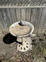 Concrete bird fountain standing in a backyard next to a wooden fence, showing the full structure with a bird figurine on top of the basin.