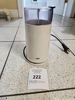 Braun coffee grinder exterior with lid on, placed on tile floor, showing cylindrical white plastic body with cord and plug.