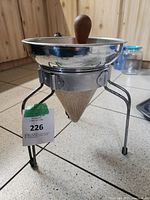 Metal canning sieve colander with conical mesh on a tripod stand with rubber feet.