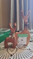 Photo showing two wooden reindeer figurines of different sizes placed on a table with a lace doily, natural wood finish and fabric ribbons visible.