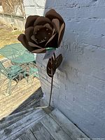 The full metal flower with stem and leaves placed outdoors against a white brick wall with a view of a patio table.