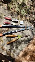 Lot of five garden tools on concrete and grass including Fiskars pruning lopper, hand trowel, hand cultivator, and pruning shears.
