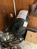 Main unit wrapped in black bag with coiled black pressure hose and manual on top, against wood panel wall.