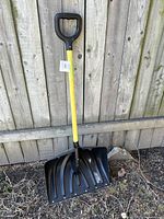 Snow shovel leaning against outdoor wooden fence on dirt ground, showing full shovel including yellow handle and black scoop.