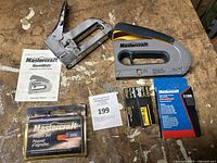 Two Mastercraft staple guns, an instruction manual, and three packages of staples including Mastercraft and Spar brands, displayed on a wooden surface.