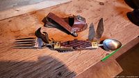 Multi-tool unfolded showing fork, spoon, knife blade with wooden handle and brass bolsters on wooden table.