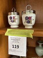 Pair of vintage Royal Worcester egg coddlers with metal lids and berry decoration on porcelain body, displayed in a wooden cabinet.