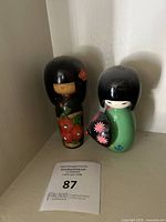 Two vintage Japanese Kokeshi dolls positioned side by side on a corner shelf. One doll wears a red floral kimono, the other green kimono with a fan.