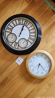 Two wall clocks on hardwood floor, one black frame with Roman numeral face and two smaller dials, the other beige frame with Arabic numerals.