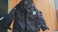 Black men's medium-size Misty Mountain waterproof jacket laid out flat on wooden floor, showing zippered pockets and adjustable cuffs.