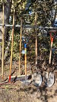 Photo showing full set of yard work tools leaning against chain link fence outdoors with trees and ground visible.