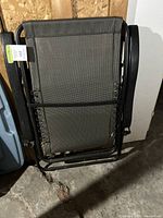 Folded zero gravity chair leaning against an unfinished wooden wall in a basement or garage setting, showing the black metal frame and grey mesh fabric seat.