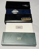 Several pen boxes stacked together showing a mix of packaging styles including black, green, and blue boxes with brand markings and a brass nameplate on one box.