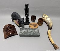 Front view of eight Inuit and indigenous art and carving pieces displayed on gray background, showing soapstone bear, wooden mask, fox figurine, tumblers, ashtray, plaque, and carved horn.