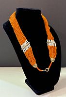 Front display of multi-strand orange bead necklace on black neck stand showing white bead accents and metal clasp