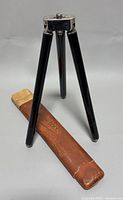 Black metal tripod standing with the original leather case laid beside it, showing tripod structure and condition.