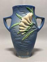 Front view of blue Roseville pottery vase with raised cream freesia flower and green leaves detail, showing wave texture background and dual handles.