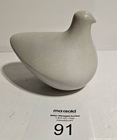 Front-left angled view of the light grey dove sculpture with rounded body and pointed tail, placed on a white surface with auction card labeled 91 visible.