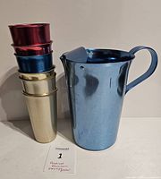 Photo of the Flamingo anodized aluminum pitcher in blue next to stacked anodized aluminum tumblers in red, blue, and gold colors