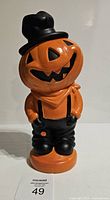 Front view of the scarecrow Halloween figurine showing pumpkin head, black top hat, orange shirt, suspenders, and pants standing on a base